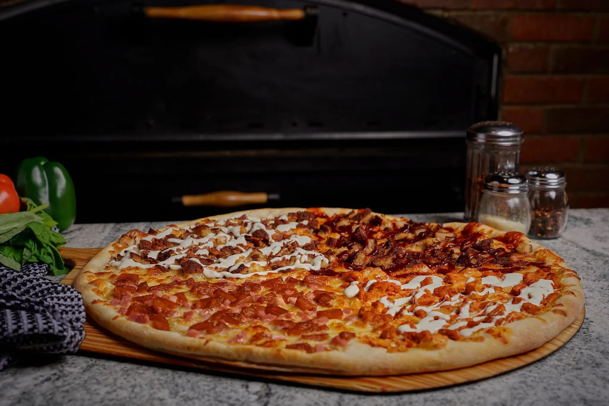 Large wood-fired pizza on a board, with peppers, spice jars, and a brick oven backdrop